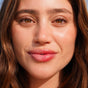 A close-up of a woman with long brown hair and brown eyes, smiling softly. A vertical dotted line highlights her center lips, tinted with FRÉ Skincares PLUMP ME Peptide Lip Tint Mineral SPF 30. Her skin is clear with minimal makeup.