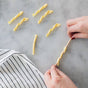 Hands wrapping pasta dough around a wooden stick and cavatelli board to form authentic Italian pasta shapes, with finished pieces and a striped cloth on a marble surface—a true FarmSteady Semolina Pasta Making Kit experience.