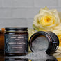 An upright jar of Chagrin Valley Soap & Salve's Activated Charcoal Clay Face Mask sits next to an open jar of gray powder, while a yellow rose in the background adds elegance to the reflective surface.