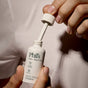 A person twists open a white bottle labeled Phil's Face and Beard Serum, featuring castor oil and marine algae, with compostable packaging details, set against a soft beige background.