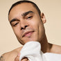 A man with short hair and a slight smile gently pats his face with a white towel after using Phil's Daily Facial Cleanser, set against a beige background.