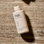 A bottle of Phil's Daily Facial Cleanser, a gentle cleanser with vitamin C, ceramides, and minerals, rests on textured stone with water droplets. The 3.4 fl. oz. (100 mL) label highlights its compostable packaging.