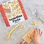 Hands shape pasta dough on a cavatelli board next to a FarmSteady Semolina Pasta Making Kit box, crafting authentic Italian pasta. The box displays assorted pasta shapes, while a striped cloth rests on the marble surface.