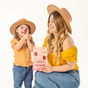A smiling woman and a young girl, both in tan hats and yellow tops, sit together. The girl holds a lollipop to her nose, while the woman holds a box of Beekeepers Naturals Kids Prebiotic Fiber Pops - Sample. Both look happy.