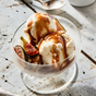 A glass bowl of vanilla ice cream topped with sliced figs and drizzled with Kosterina Fig Balsamic Vinegar sits on a rustic white wooden surface.