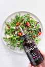 A hand holds a bottle of Kosterina Fig Balsamic Vinegar above an arugula salad topped with apples, walnuts, and a drizzle of balsamic vinegar on a white surface.
