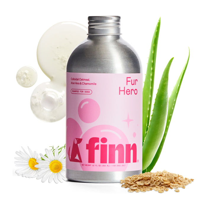 A silver and pink Finn Fur Hero Shampoo for Dogs bottle sits with chamomile, aloe vera, oatmeal, and creamy drops. This all-natural shampoo soothes sensitive canine skin using colloidal oatmeal, aloe vera, and chamomile.