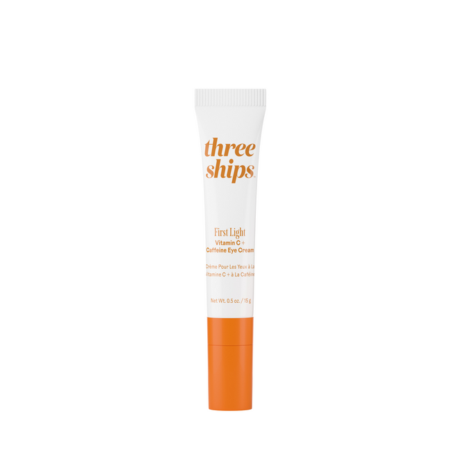 A tube of Three Ships First Light Vitamin C + Caffeine Eye Cream, with an orange cap and label, stands upright against a white background.