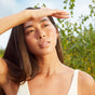 A woman with long hair stands outdoors among green foliage, wearing a white top and delicate necklace. She applies Three Ships First Light Vitamin C + Caffeine Eye Cream under her eyes, brightening her skin as she shields her eyes from the sun.