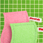 A pink and a light green Floofah Luffa + Wood Dual Kitchen & Dish sponge with labels attached, positioned against a green tiled background.