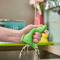 A hand squeezes a wet, green Luffa + Wood Dual Kitchen & Dish Sponge under a running faucet.