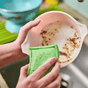 Hands cleaning a dirty bowl with a green dual-sided Floofah Luffa + Wood Dual Kitchen & Dish Sponge.