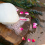 A white Floofah Exfoliating Shower Loofah with soap suds rests on a wooden board, surrounded by scattered pink rose petals and green sprigs.