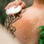 A person with a large floral tattoo on their shoulder washes their back with the Floofah Exfoliating Shower Loofah, creating bubbles and soap suds on their skin for a touch of sustainable cleaning.