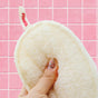 A hand with glittery nails holds a beige Exfoliating Shower Loofah made from plant-based luffa gourd. A tag on the loofah reads Floofah. The background is a pink tiled wall.