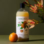A bottle of Koala Eco Floor Cleaner Refill, featuring a botanical illustration on the label, stands next to a fresh mandarin and a sprig of flowers, set against a green surface and wall.