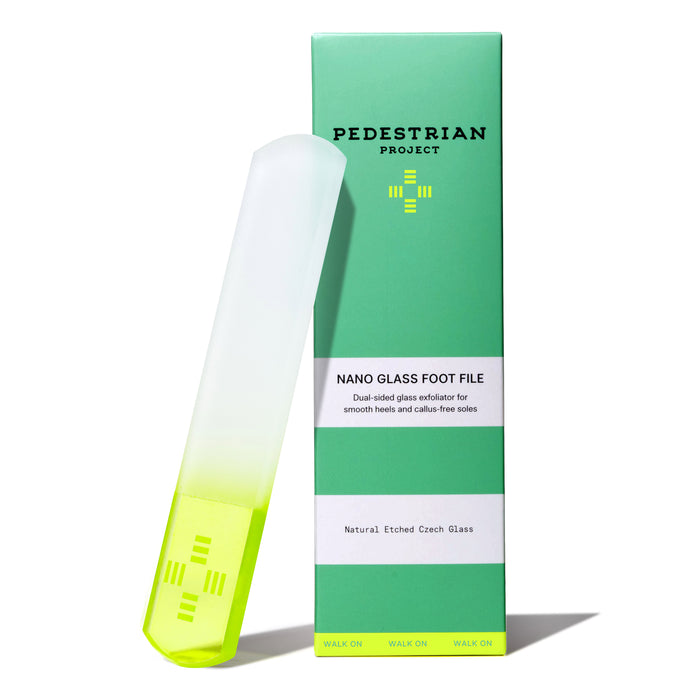 The Nano Glass Foot File by Pedestrian Project, featuring a frosted handle with neon green tip, stands upright beside its green and white box labeled for smooth heels and gentle callus removal.