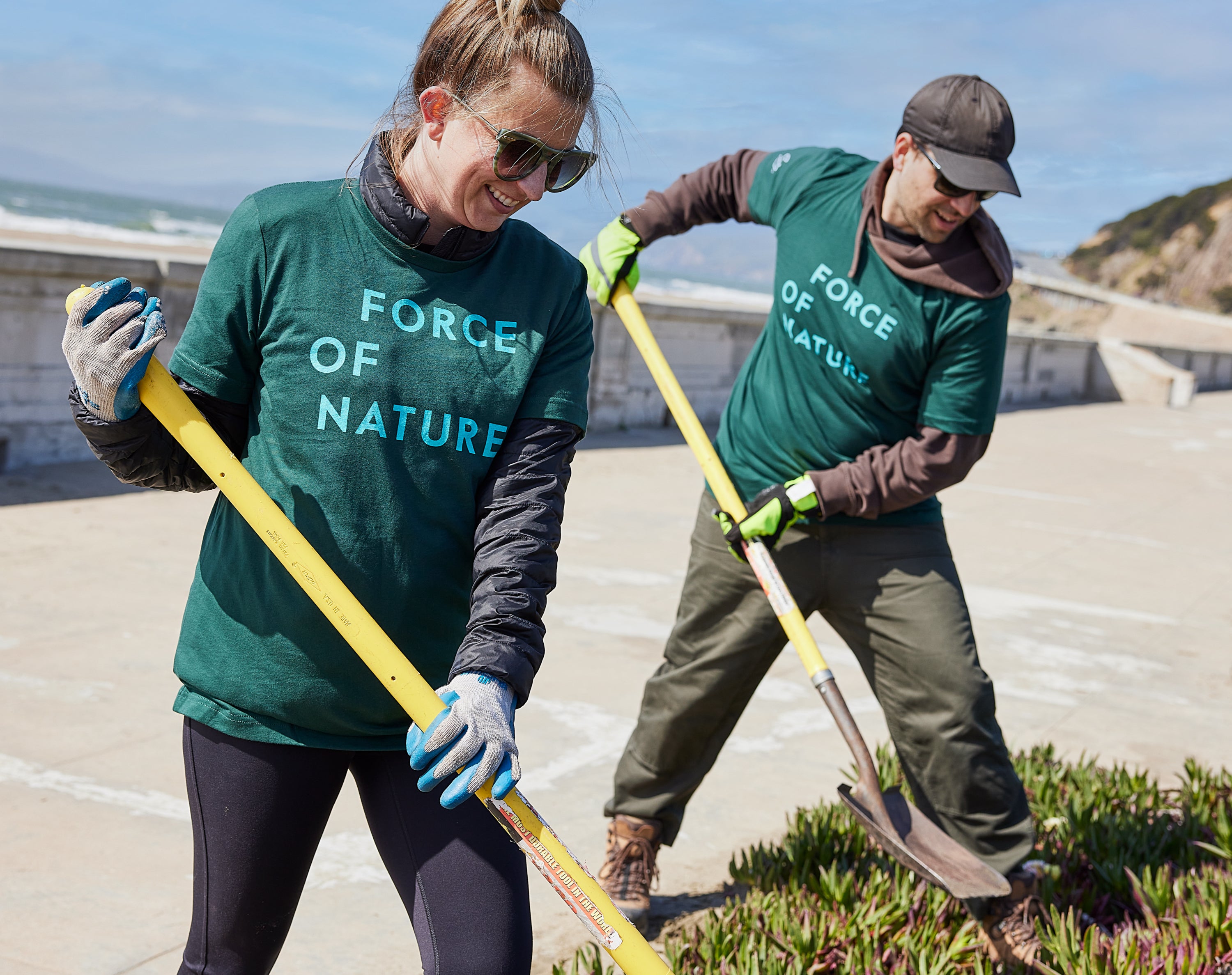 Two people wearing green Force of Nature shirts and gloves use tools to tend to plants outdoors on a sunny day, near a concrete walkway and coastline.