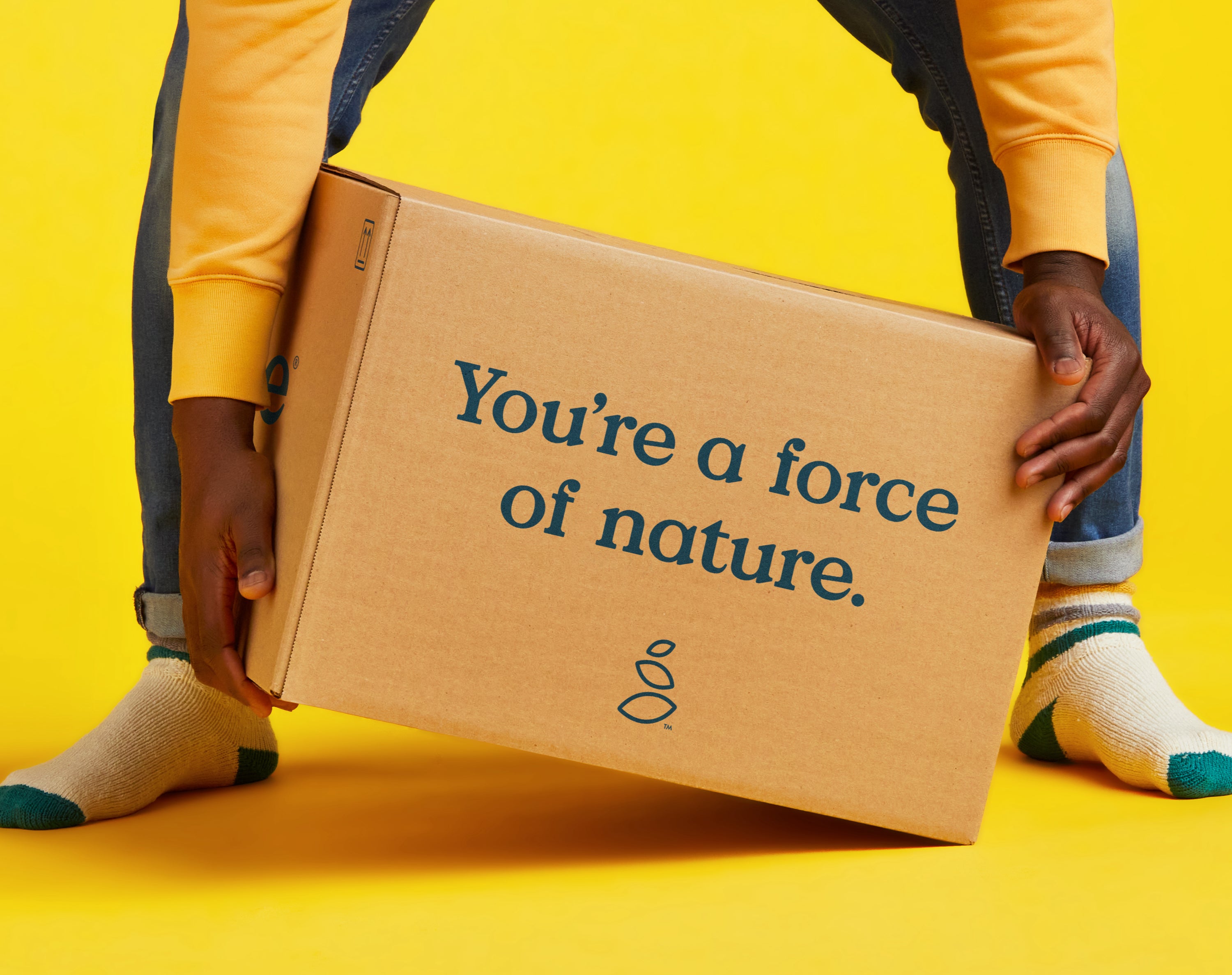A person wearing a yellow sweater and colorful socks bends down to lift a cardboard box with the words Youre a force of nature. printed on it, against a bright yellow background.