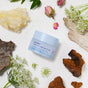 A light blue jar of Ursa Major Forest Alchemy Eye Cream, designed to target fine lines and dark circles, sits on a white surface surrounded by dried flowers, tree bark, and white blossoms in a botanical arrangement.