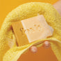 A hand holds a foamy yellow towel and a cream-and-orange Foxly Moisturizing Bar Soap, its name carved on the bar, set against a bright yellow background—highlighting nourishing handcrafted soaps.