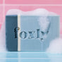 Foxly Moisturizing Bar Soap, a blue and white handcrafted bar with "Foxly" etched on it, rests foamy on a wet surface before pink tiles—crafted from clean ingredients and nourishing oils for a gentle cleanse.