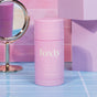 A pink stick of Foxly Deodorant stands on blue tiles with a round mirror and pastel geometric shapes in the background, offering 24-hour odor protection and an aluminum-free formula.