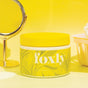 A yellow jar of Foxly Body Scrub in Citrus Beach sits on a yellow surface. A round mirror and a yellow basket with a bath sponge are in the background, all set against a bright yellow backdrop.