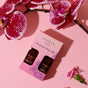 The Foria Intimacy Passion Play Kit with Organic Botanicals features two small brown bottles—Massage Oil and Awaken Arousal Oil—packaged in a pink and purple box, shown on a pink surface with purple orchids and a pink carnation beside it.