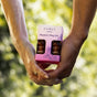 Two people gently hold hands while showcasing the Foria Intimacy Passion Play Kit with Organic Botanicals—featuring arousal and massage oils—set against a blurred, sunlit outdoor backdrop with lush green foliage.