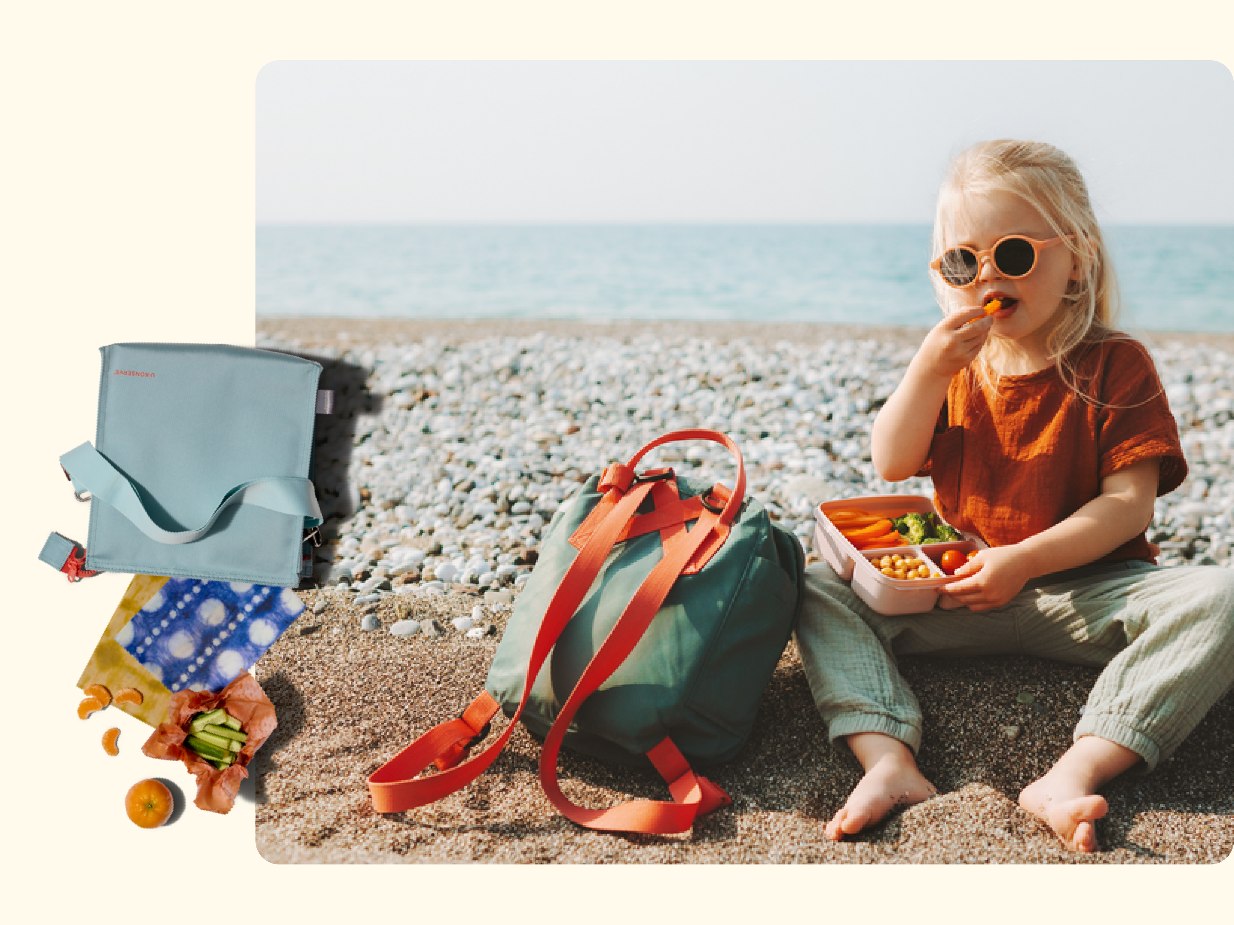 A young child wearing sunglasses sits on a pebble beach eating from a lunchbox. Nearby are a green backpack, a light blue cooler bag, and a colorful cloth with snacks on it. The sea is visible in the background.