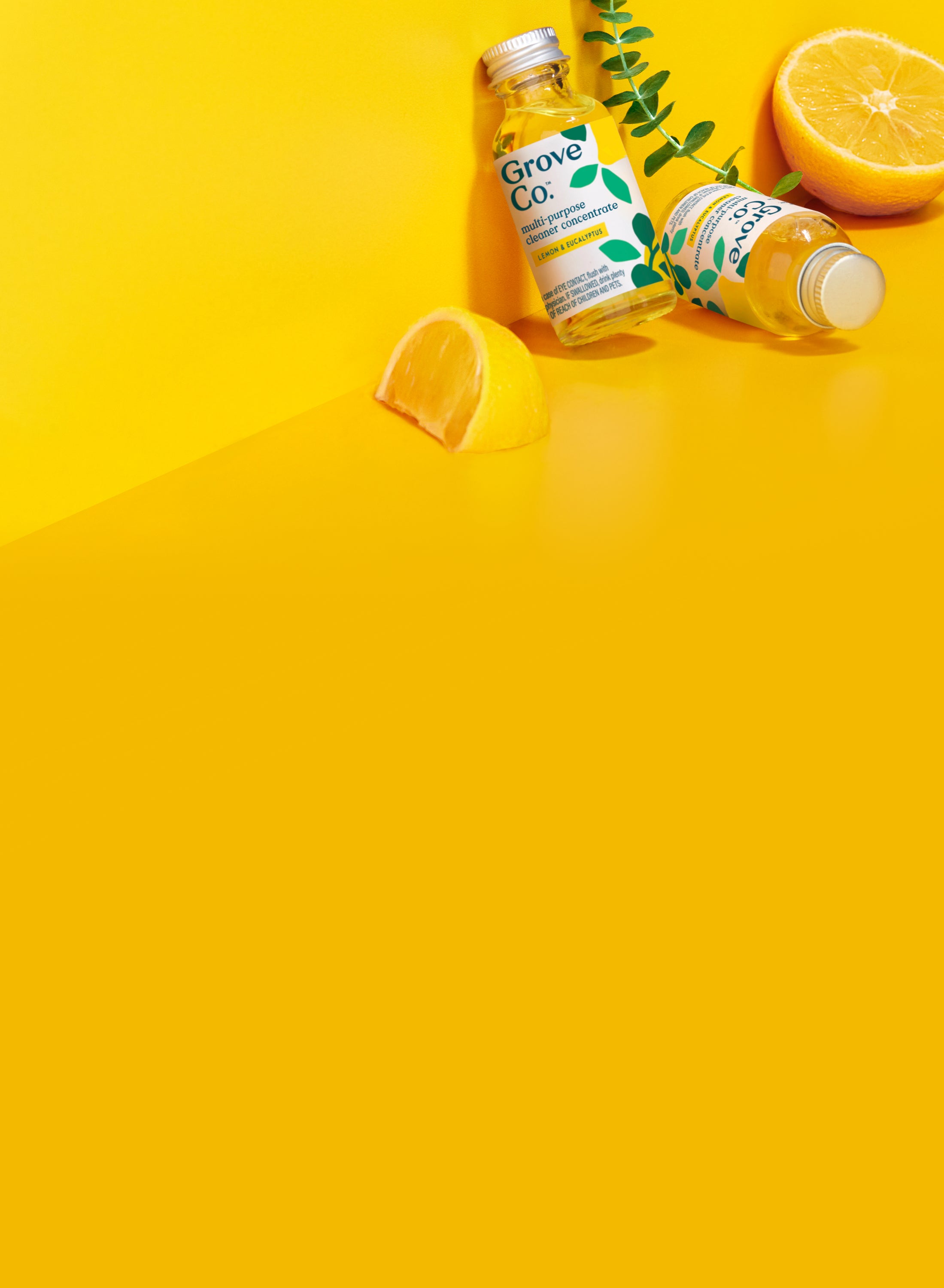 Two Grove Co. cleaning bottles and fresh lemon slices are arranged on a bright yellow background with a small green leaf accent.