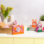 A bright, organized kitchen counter displays the Grove Co. Multi-Purpose Cleaner Starter Set - Summer Collection with plant-based ingredients, plates, a reusable spray bottle, towels in a basket, a potted succulent, and fresh flowers against tiled walls.