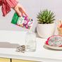 Someone pours Grove Co. Hand Soap Refill - Summer Collection from a vibrant carton into a glass dispenser on the counter, with a potted succulent, cut dragon fruit, and a basket in the background.