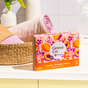 A colorful box of Grove Co. Laundry Detergent Sheet Set - Summer Collection sits on a counter next to a woven basket with a rolled pink towel, with cleaning supplies and plants visible in the background.