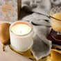 A Grove Co. Soy Wax Blend Candle from the Winter Collection in a ribbed glass holder sits on a gold tray beside a ripe pear, a light gray cloth, a glass of yellow drink, and a Grove Co. box in the background.