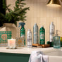 A bathroom counter with Grove Co. cleaning products, hand soap, a Grove Co. Soy Wax Blend Candle - Winter Collection, a glass soap dispenser, a folded towel, and a potted evergreen branch against cream-colored tile walls.