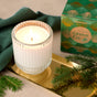 A lit Grove Co. Soy Wax Blend Candle from the Winter Collection in a ribbed glass holder sits on a gold tray beside pine branches, a green cloth, and the festive green and gold Grove Co. box, creating a cozy holiday vibe.