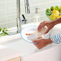 A person uses Grove Co. All-Purpose Coconut Scrubber Sponges to clean a white plate with food stains under a kitchen sink faucet, while a soap dispenser and a bowl of lemons and limes sit in the background.