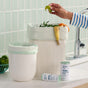 A person drops food scraps into a white compost bin lined with Grove Co. Compostable Food Scrap Bags, next to a smaller bin and two rolls of Grove Co. small bin bags on the kitchen counter.