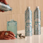 A person pours Grove Co. Hand Soap Refill - Winter Collection from an aluminum bottle into a ribbed glass dispenser, while another Grove Co. dispenser and plant-based hand soap with natural origin fragrance sit on the countertop.