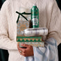 A person in a white cable-knit sweater holds stacked Grove Co. gift boxes, metal bottles, a Hand Soap Refill - Winter Collection, a candle with a green ribbon, and a blue patterned cloth. The holiday packaging has festive green and gold designs.