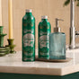 A Grove Co. Hand Soap Refill - Winter Collection bottle stands next to a ribbed blue glass dispenser on a white bathroom countertop, with a candle and green decor in the background.