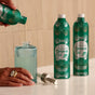 A person pours liquid from a green Grove Co. Hand Soap Refill - Winter Collection bottle into a ribbed glass dispenser, with another Grove Co. hand soap on the countertop nearby.