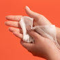 A person cleans their left hand with a Grove Co. Hand Sanitizing Wipe—Kills 99.9% of Germs—against a solid orange background.
