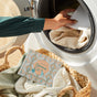 A person adds Grove Co. Laundry Detergent Sheets - Winter Collection to a front-loading washer filled with clothes, while a woven laundry basket and the Grove Co. detergent sheets box sit nearby.