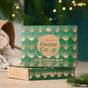 A box of Grove Co. Home Staples Set - Winter Collection with green and gold art deco design sits on a light surface, softly lit with blurred holiday lights in the background.