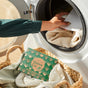 A hand places laundry into a washing machine. In the foreground, a basket holds clothes and Grove Co. Laundry Detergent Sheets Set – Winter Collection for naturally fresh, clean laundry every time.