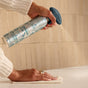 A person sprays Grove Co. Multi-Purpose Cleaner - Winter Collection from a decorative bottle onto a surface, wiping with a cloth in front of beige tiles.