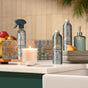 A kitchen counter displays Grove Co. Multi-Purpose Cleaner - Winter Collection, a soap dispenser, stacked dishes with a pear and apples, a lit candle, decorative boxes, and greenery above against a tiled wall.